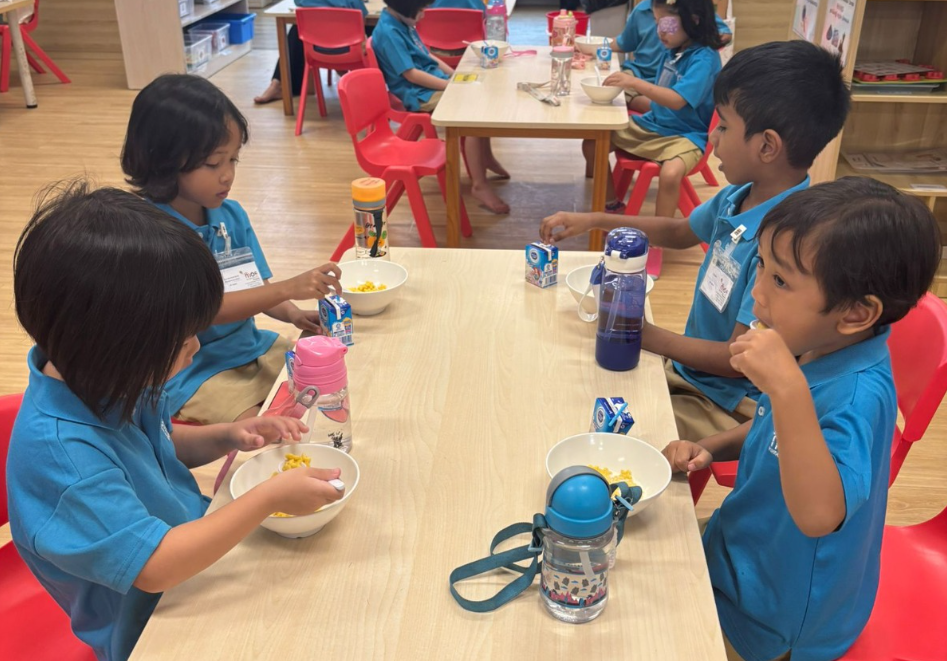 Children in blue uniforms sit at tables eating from bowls, with milk cartons and water bottles in a classroom.